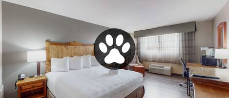 A hotel room with a large bed, a desk, a chair, a nightstand, a lamp, a telephone, and a window with curtains.