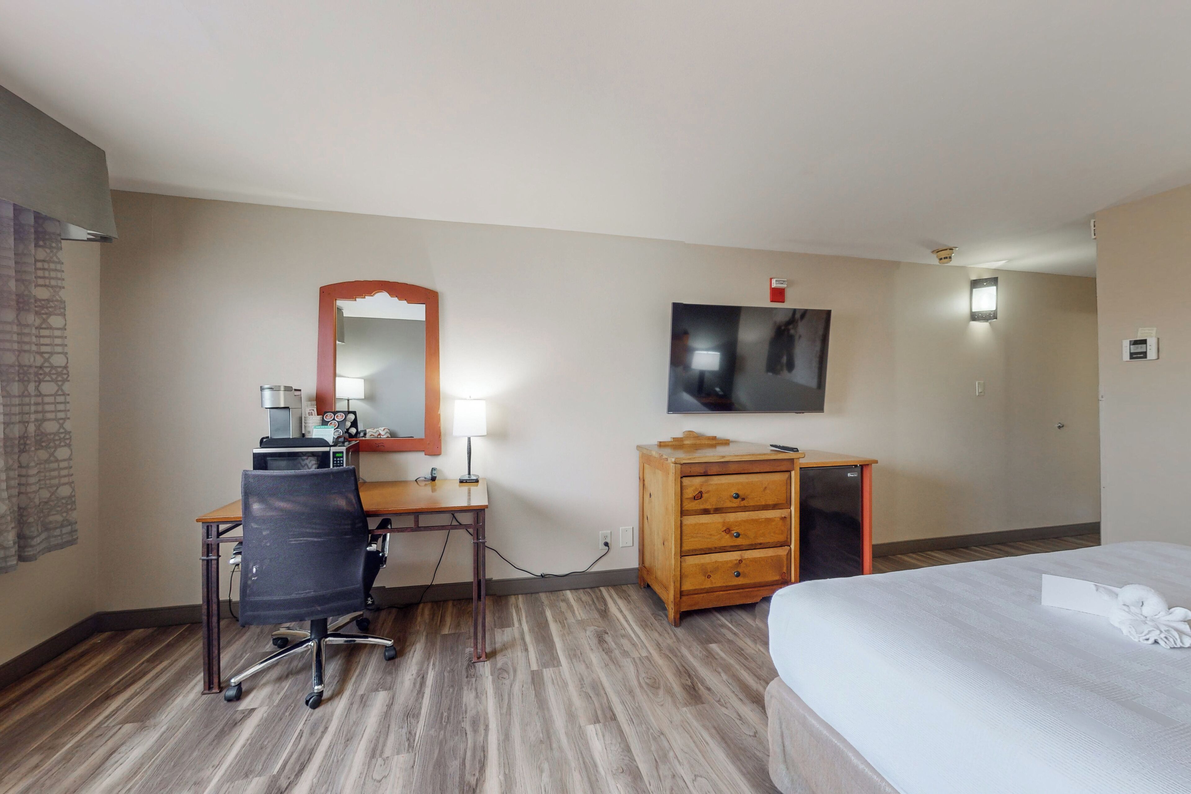 Ein Hotelzimmer mit Schreibtisch aus Holz, einem Sessel, einer Kommode, einem Bett, einem flachbildfernseher an der Wand und einem kleinen Tisch mit Lampe.