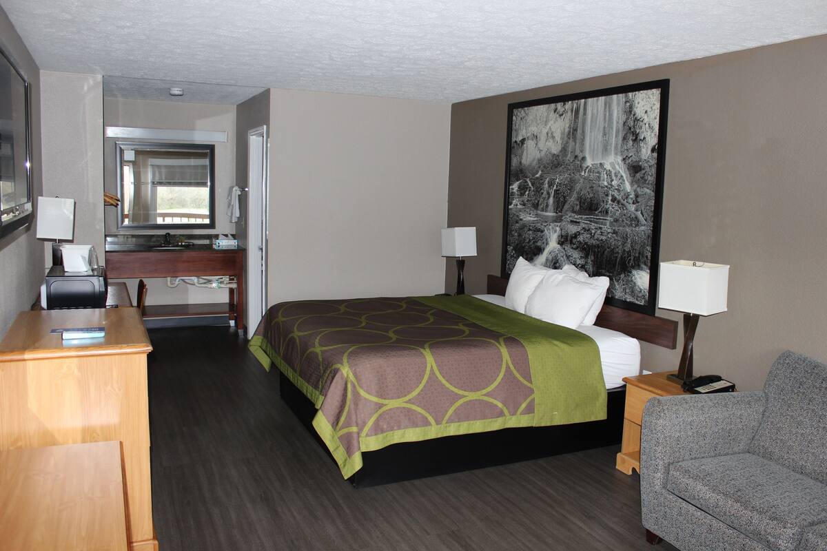 Habitación de hotel con cama, escritorio, silla, mesita de noche, lámpara, un cuadro y baño con espejo y lavabo.