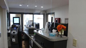 Zona de recepción de un hotel con un cajero automático, una computadora y un mostrador con un jarrón de flores.