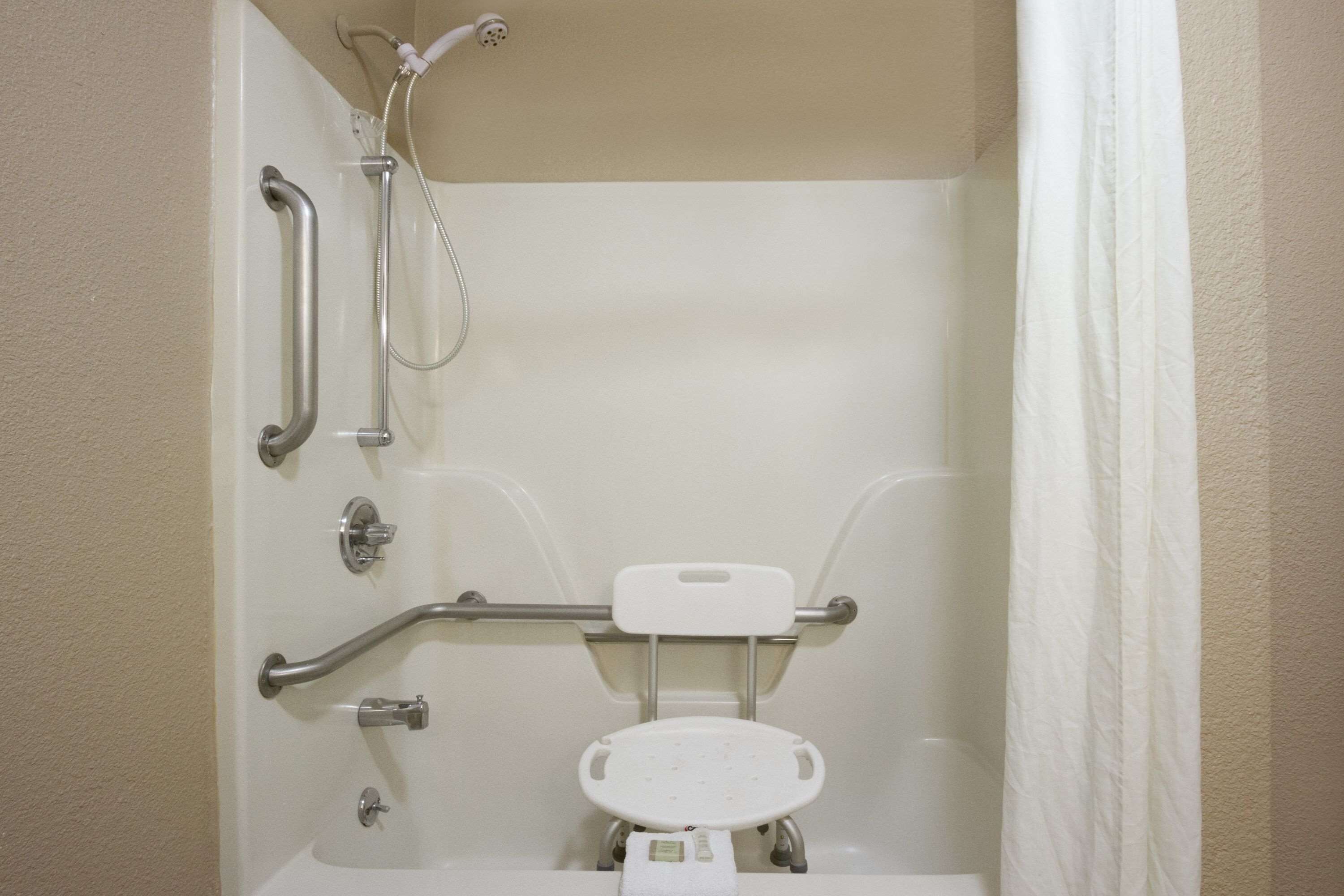Un baño con una bañera blanca, barras de apoyo, un cabezal de ducha manual y un inodoro blanco con asiento de seguridad.