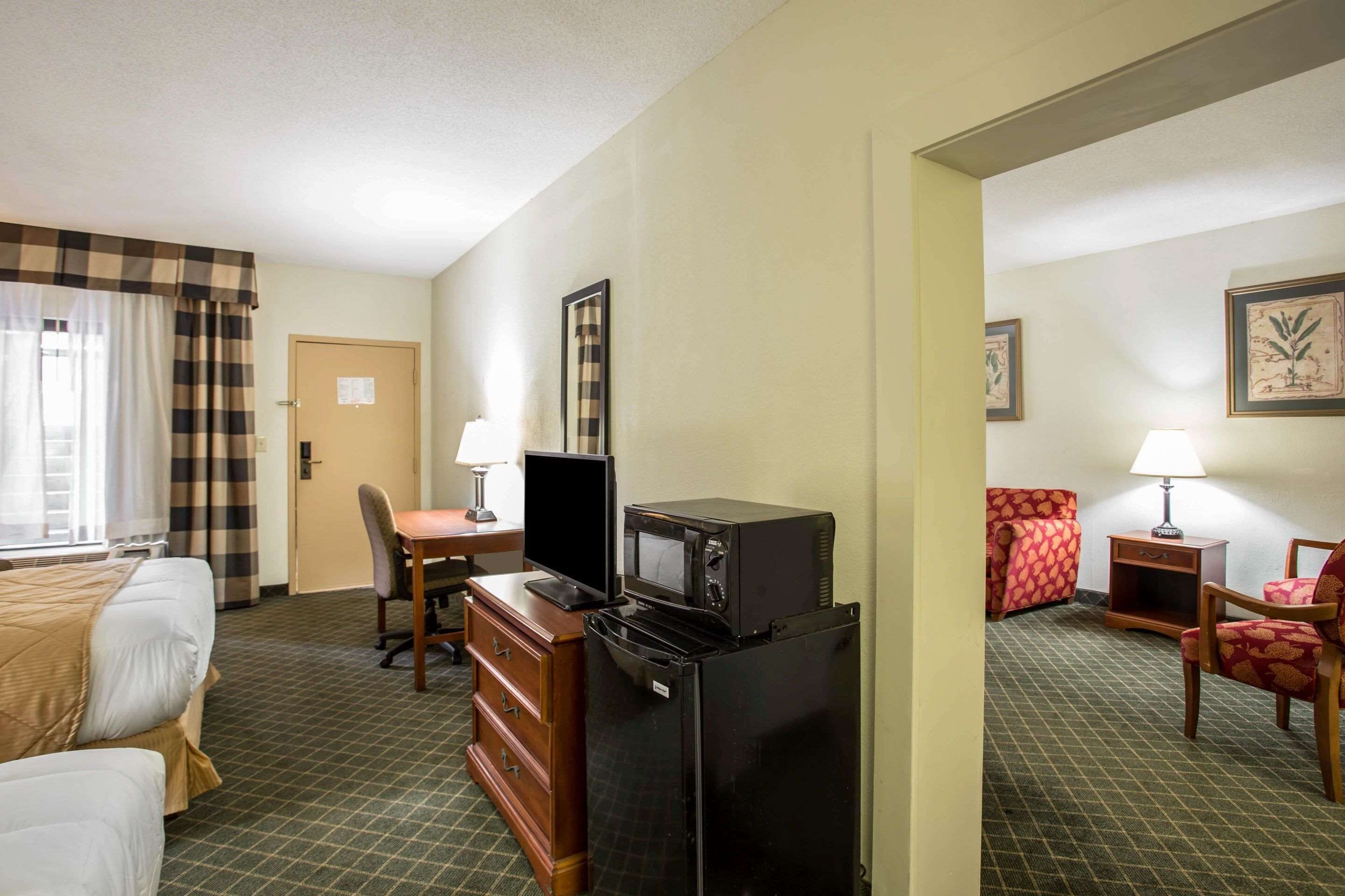 Habitación de hotel con cama, escritorio, computadora, refrigerador, microondas, silla y lámpara.