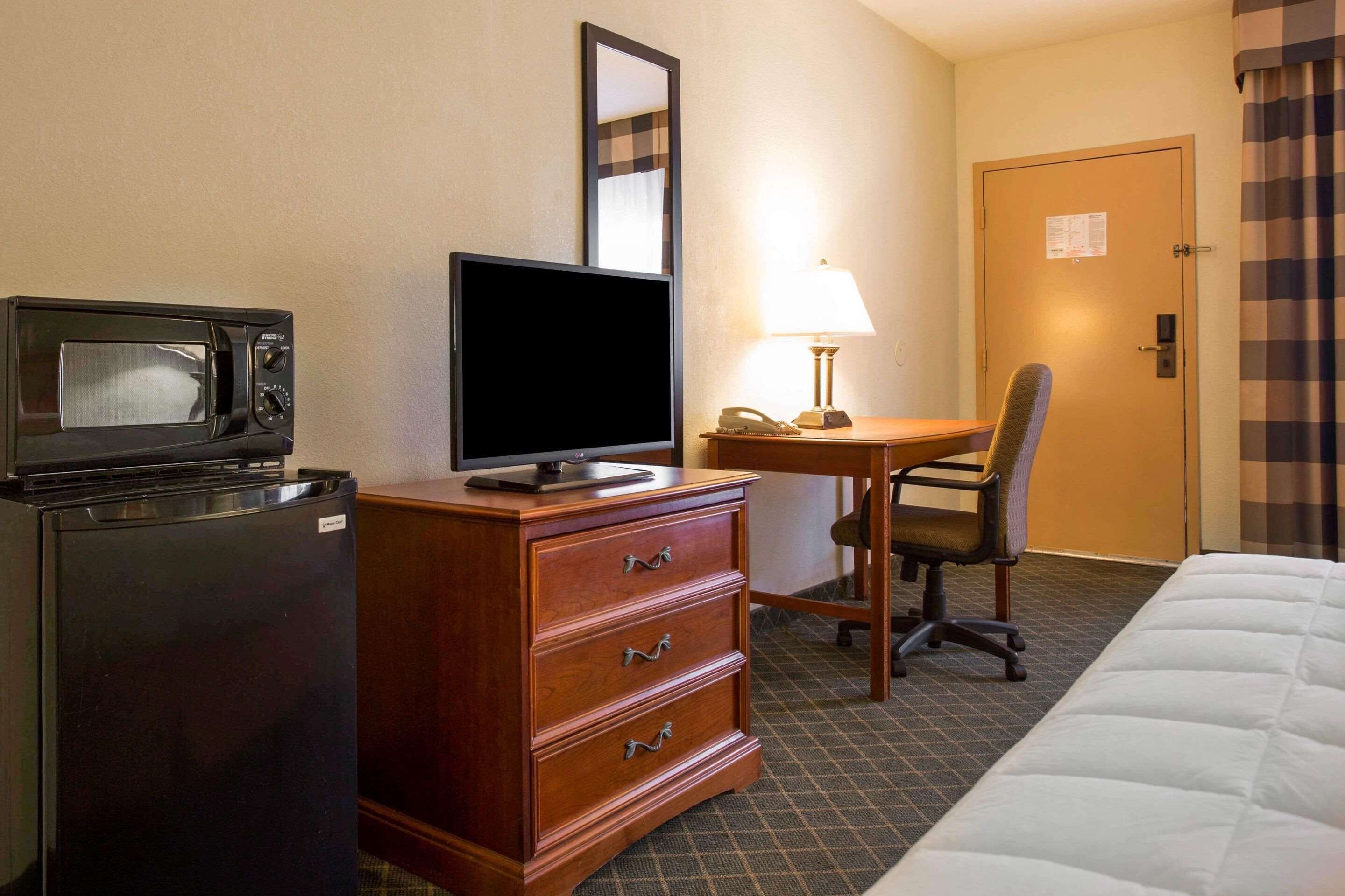A hotel room with a bed, a desk with a chair, a microwave, a refrigerator, and a lamp.