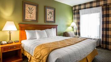 A neatly made bed with a white comforter and a mustard-colored throw blanket. There are two bedside tables, each with a lamp and a phone. The room has green walls and two framed botanical prints above the bed.