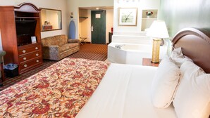 Quarto de hotel com uma cama grande, cómoda de madeira, televisão, sofá, cadeira, banheiro com banheira e um espelho na parede.