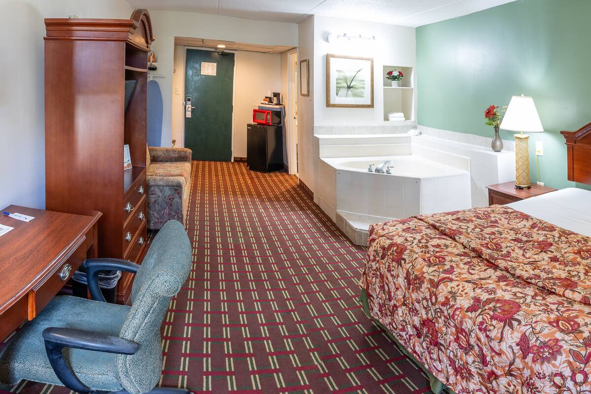 A hotel room with a bed, a desk, a chair, a small refrigerator, a sink, and a mirror.