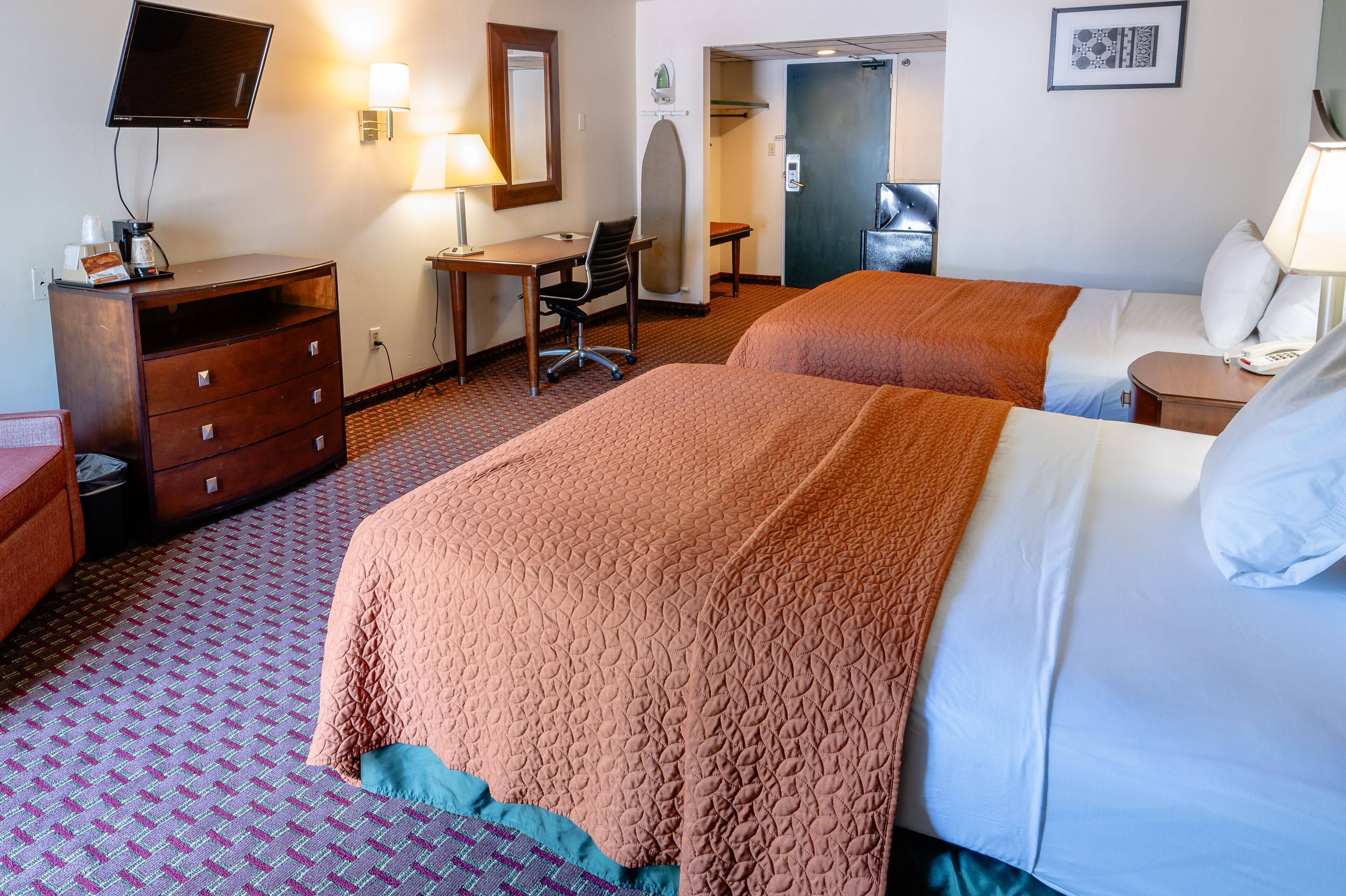 A hotel room with two beds, a desk, a chair, a dresser, a TV, and a bathroom door.