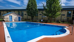 Piscina retangular com borda branca, rodeada por um revestimento vermelho, e um pequeno edifício com porta azul e placa de boia salva-vidas.