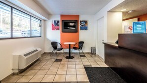 A reception area with a reception desk, two chairs, a small table, a TV, and a window with a view of parked cars. - Motel 6 Birmingham, AL (Birmingham)