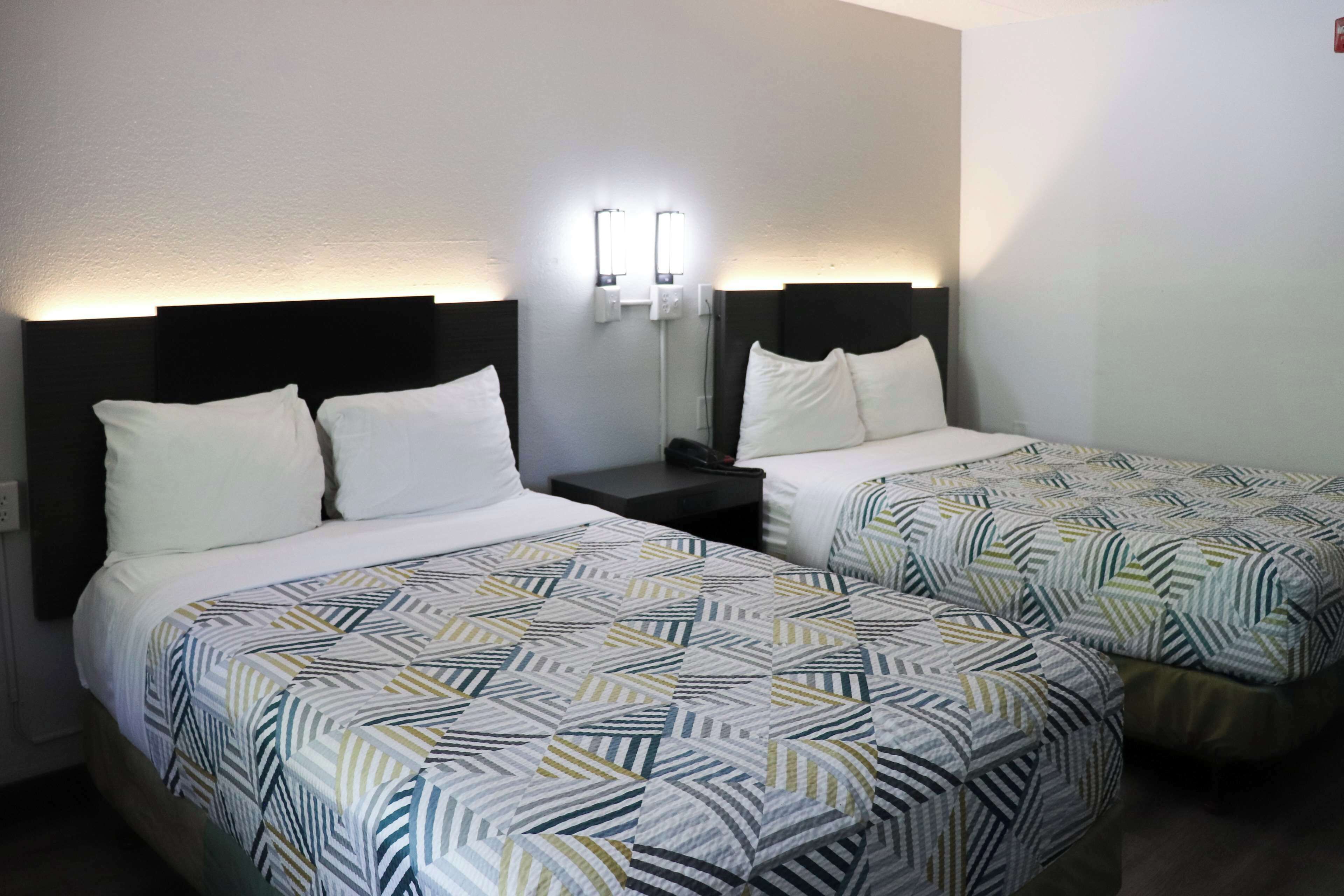 A hotel room with two beds, each with a patterned bedspread, a nightstand with a lamp, and wall-mounted lights.