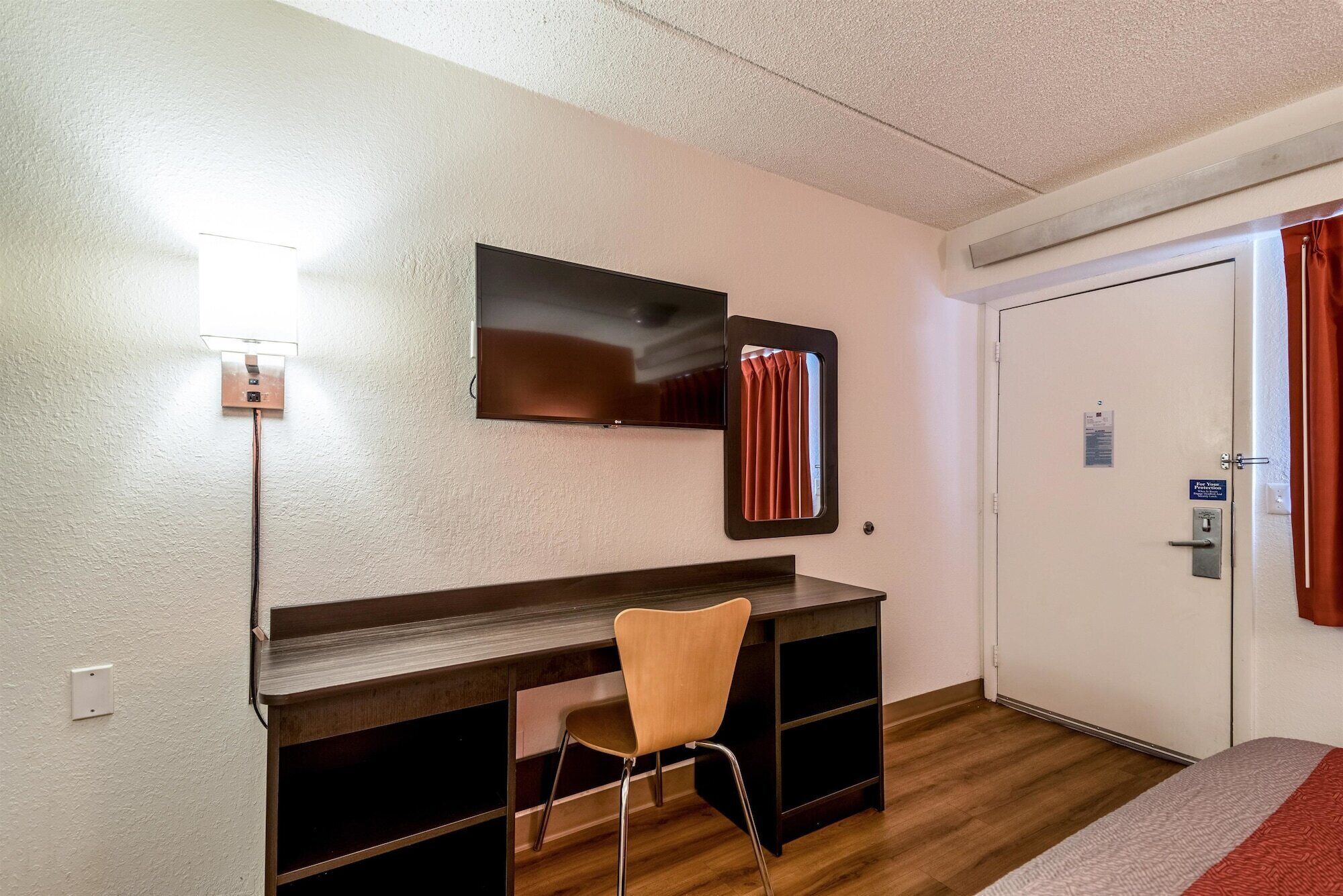 Ein Hotelzimmer mit einem Holztisch, einem Sessel, einem flachbildfernseher an der Wand, einer Tür mit einem Schild und einem kleinen Fenster mit Vorhängen.