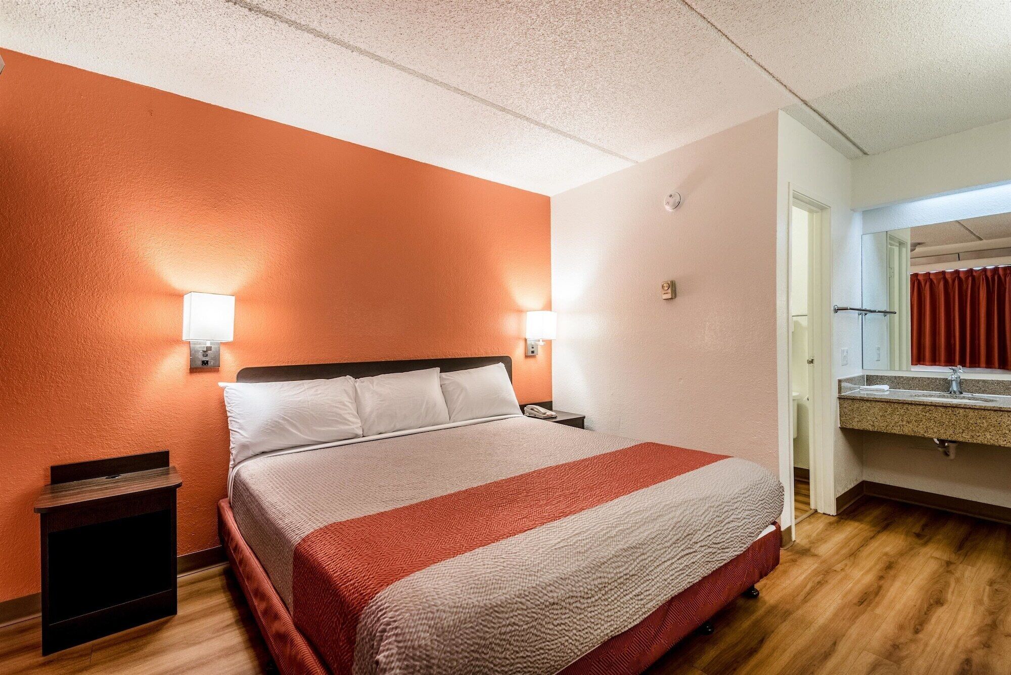 A hotel room with a bed, bedside lamps, a nightstand, a TV, and a bathroom with a mirror and sink.