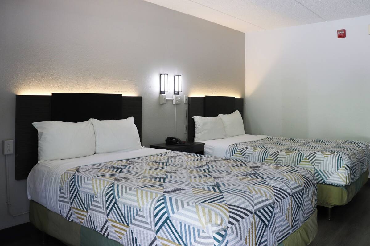 A hotel room with two beds, each with a patterned bedspread and white pillows. There are bedside tables with lamps and a red emergency exit sign on the wall.