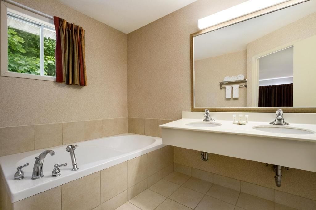 Un baño con bañera, dos lavamanos, un espejo grande y una ventana con cortinas.
