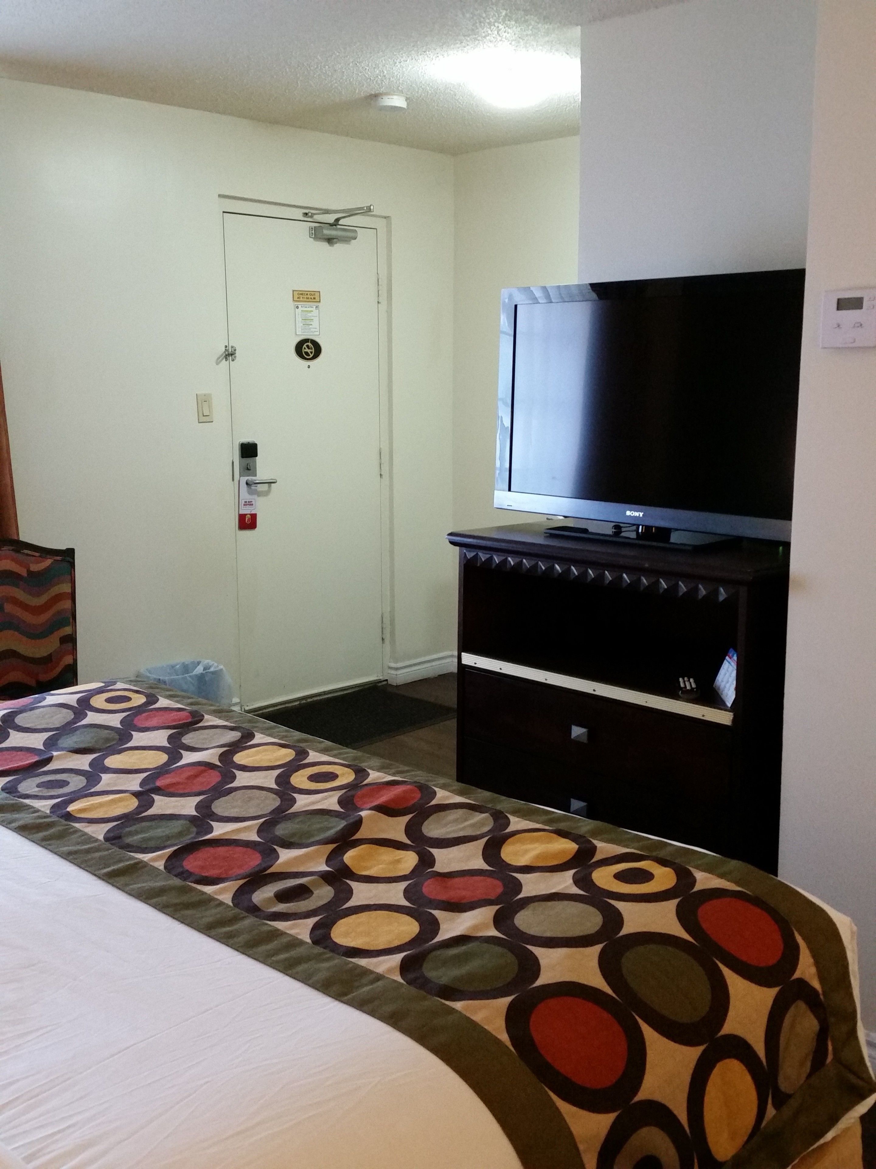 A hotel room with a bed, a television on a stand, a door with a security lock, and a patterned bedspread.