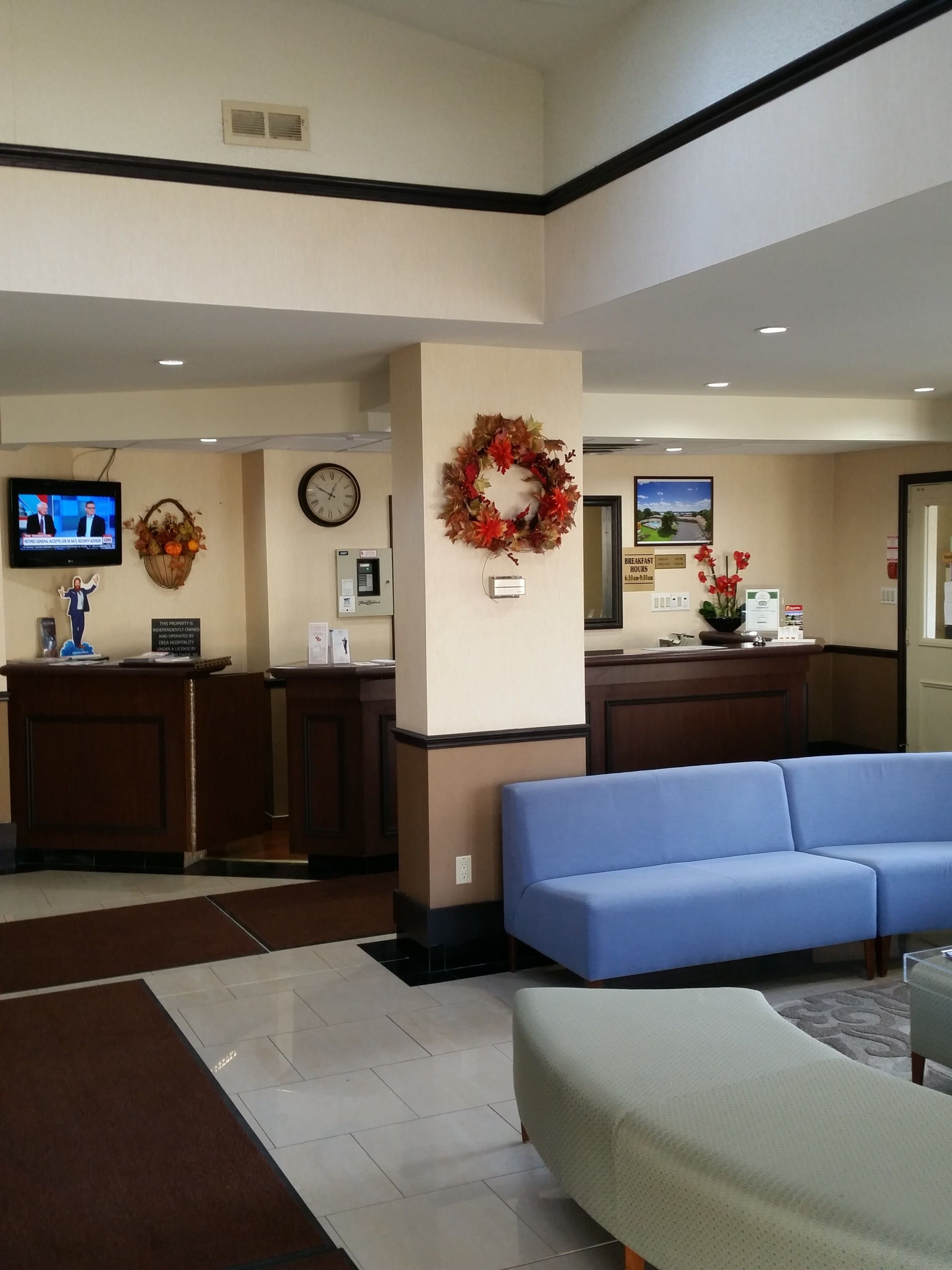 Un hall de hotel con mostrador de recepción, un sofá azul, un reloj y un televisor que muestra noticias.