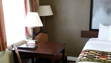 A hotel room with a bed, a desk with a chair, a lamp, and a window with curtains.