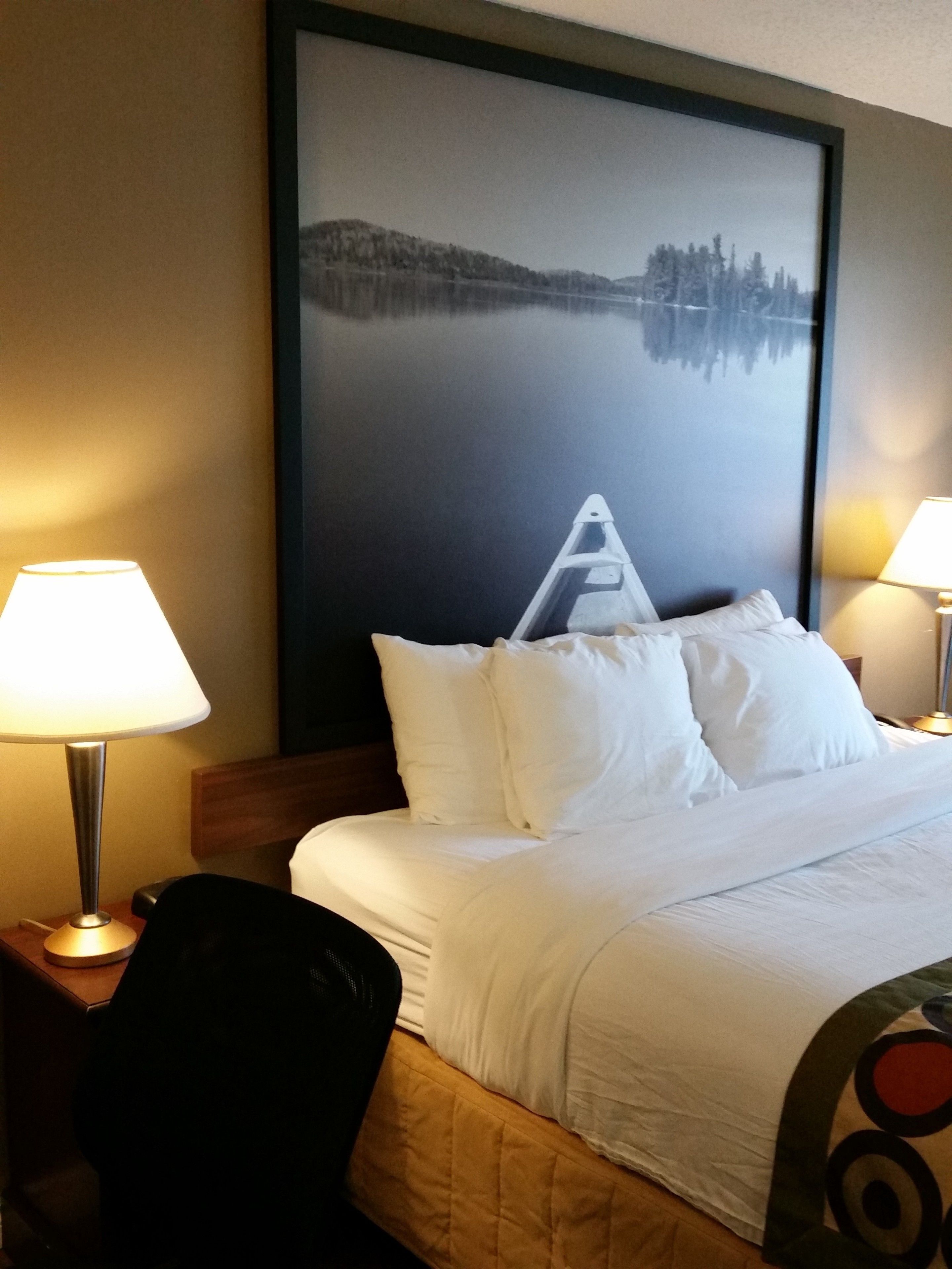 Una habitación de hotel con una cama, dos lámparas de noche, una silla y un cuadro grande enmarcado en la pared.