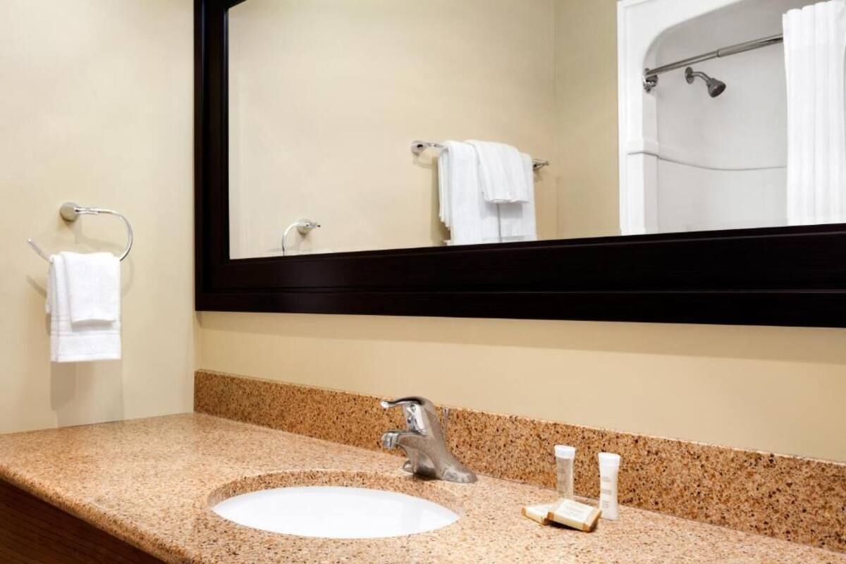 Un baño con encimera de granito, un lavabo blanco, un espejo y una ducha visible en el reflejo.