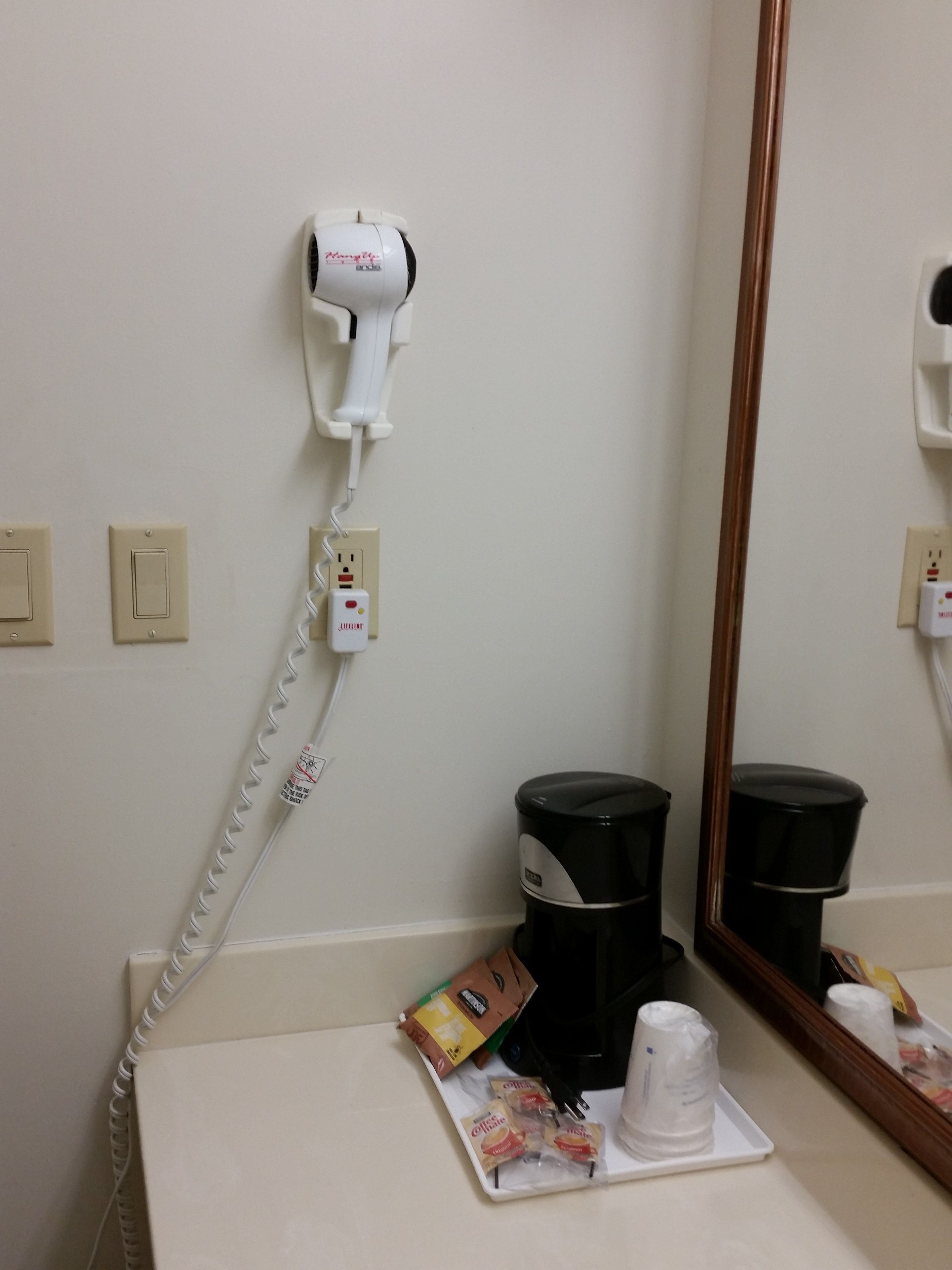 A hotel room with a hairdryer on a wall socket, a coffee maker, and a tray with coffee packets and a cup.