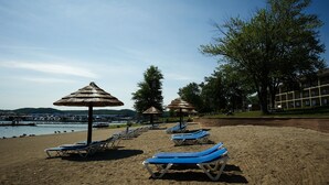 A beach with sun loungers and thatched umbrellas, a building in the background, and a body of water. - Fourwinds Lakeside Inn & Marina (Bloomington)