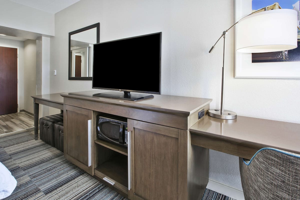 A hotel room with a wooden desk, a flat-screen TV, a microwave, a lamp, and a framed picture on the wall.