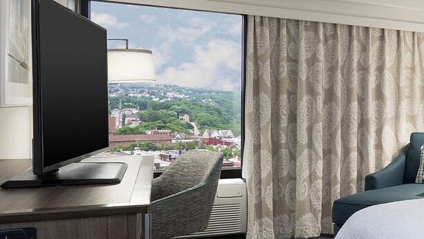 Ein Hotelzimmer mit großem Fenster, Flachbildfernseher, Schreibtisch, Stuhl und Bett.