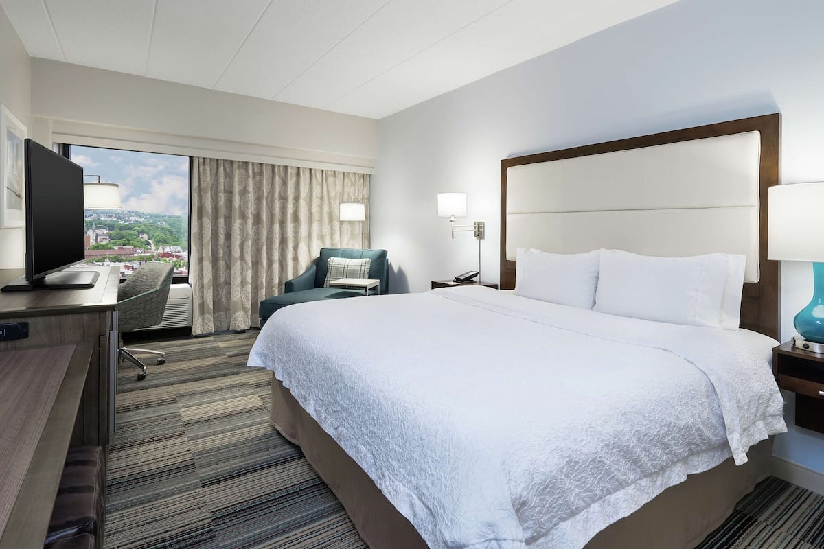 A hotel room with a large bed, a desk with a TV, a chair, and a view of the city.