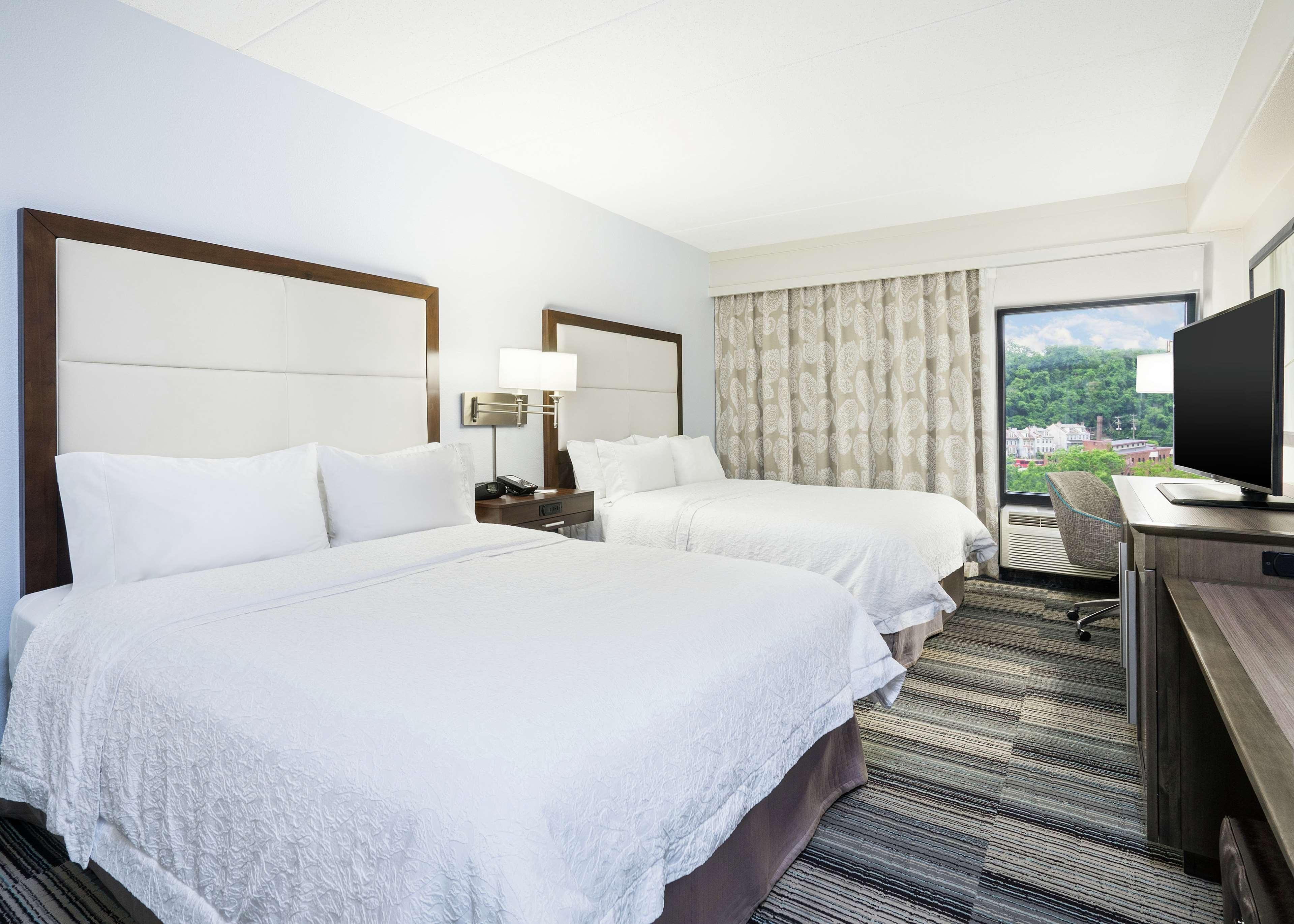 Habitación de hotel con dos camas, un escritorio con televisor, una ventana con vista a la ciudad y una mesita de noche con lámpara.