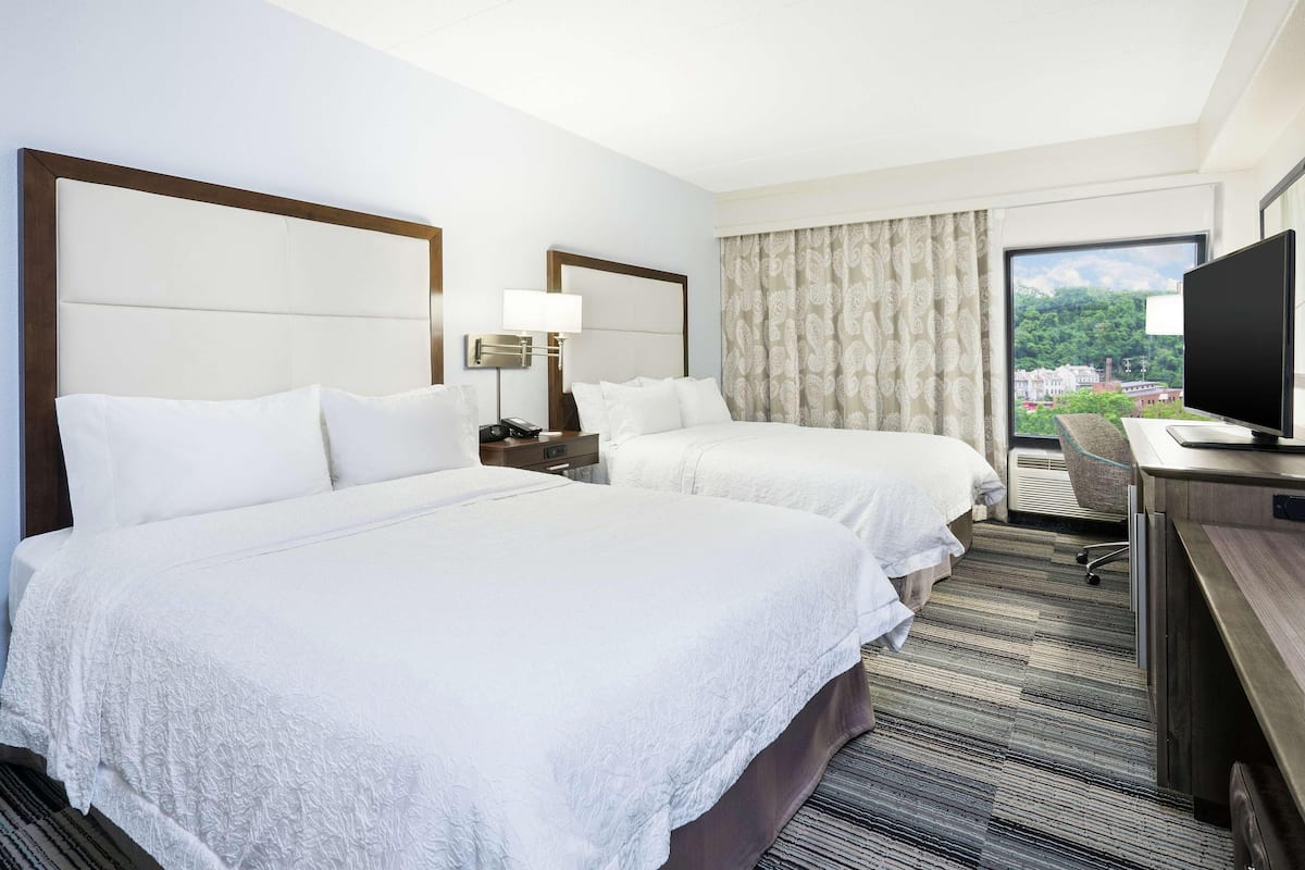 A hotel room with two beds, a desk with a television, a window with a city view, and a nightstand with a lamp.