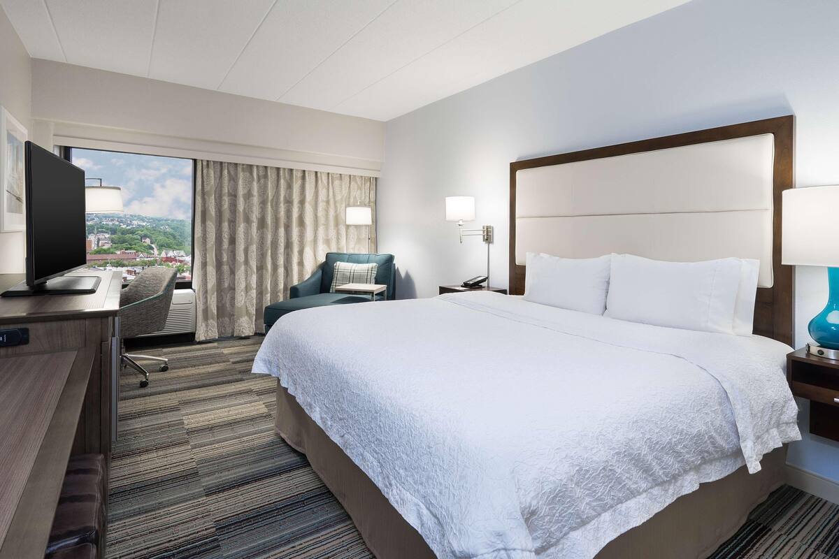 Habitación de hotel con una cama grande, un escritorio con televisor, una silla, una mesita de noche con lámpara y vistas a la ciudad por la ventana.