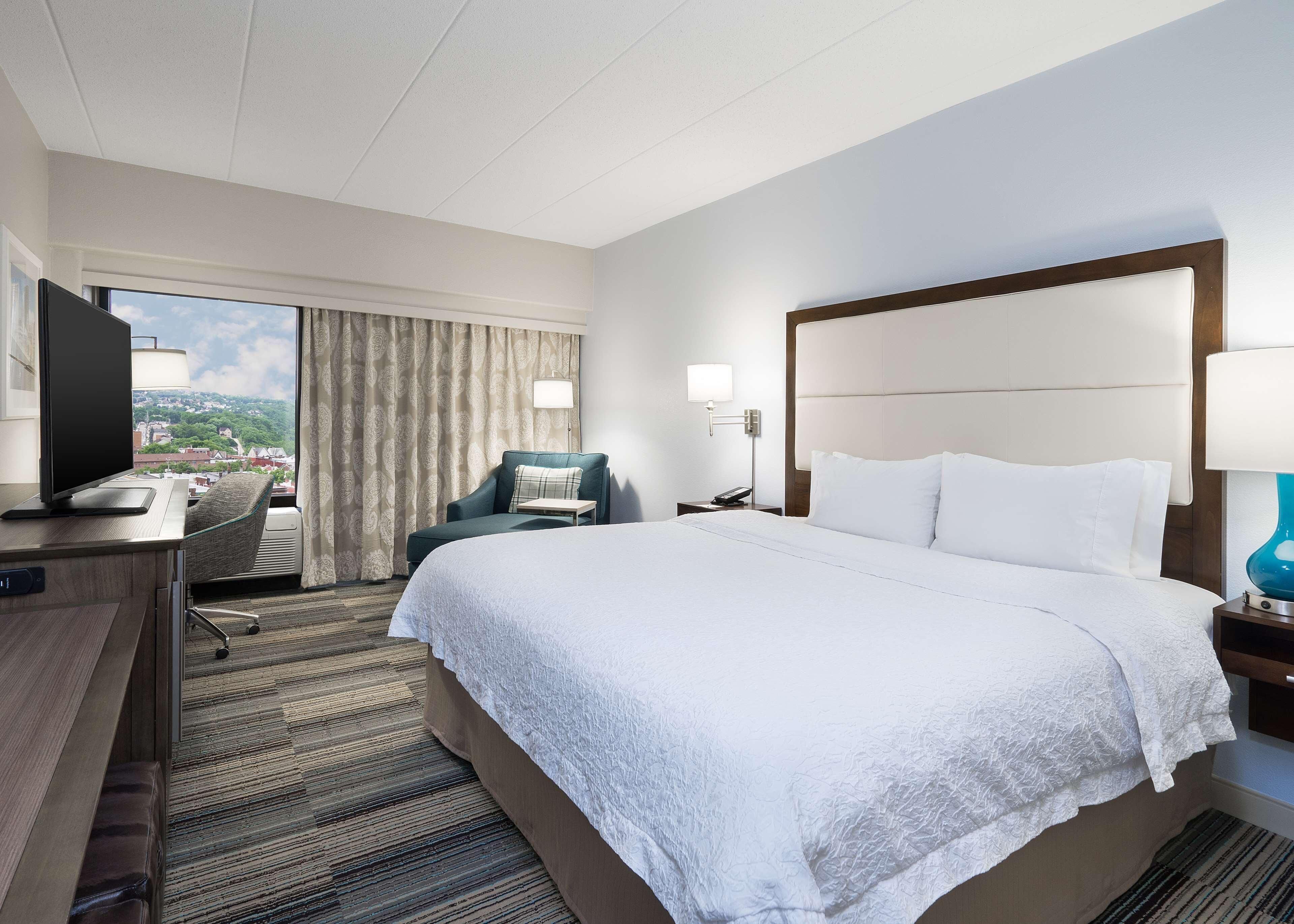 Habitación de hotel con una cama grande, un escritorio con televisor, una silla, una mesita de noche con lámpara y vistas a la ciudad por la ventana.