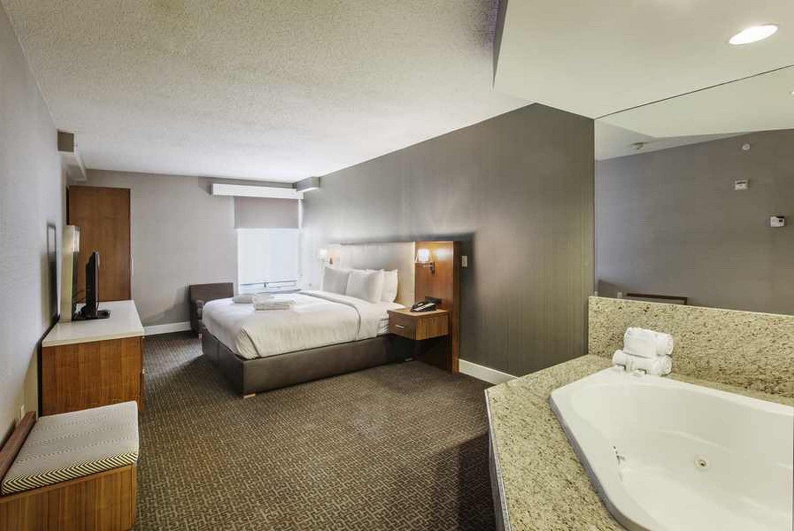 A hotel room with a large bed, a desk, a chair, a bedside table, a nightstand, a bathroom with a bathtub, and a mirror.