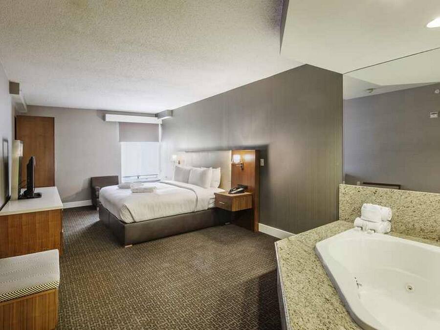 A hotel room with a large bed, a desk, a chair, a bedside table, a nightstand, a bathroom with a bathtub, and a mirror.