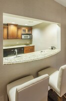 Una cocina con armarios de madera, microondas y encimera de granito. Hay dos sillas blancas frente al mostrador.