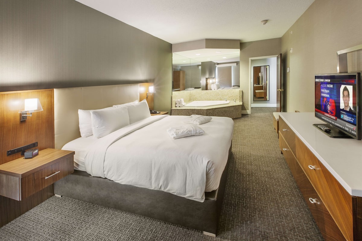 A hotel room with a large bed, a bedside table with a lamp, a TV, and a bathroom with a mirror and sink.