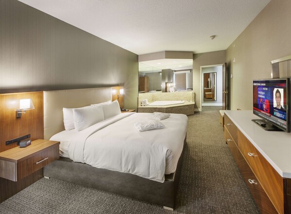 A hotel room with a large bed, a bedside table with a lamp, a TV, and a bathroom with a mirror and sink.