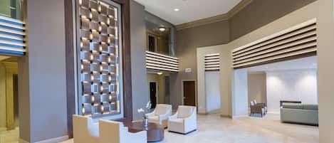 A spacious lobby with a modern design, featuring a seating area with sofas and armchairs, a decorative wall panel, and a large window providing natural light.