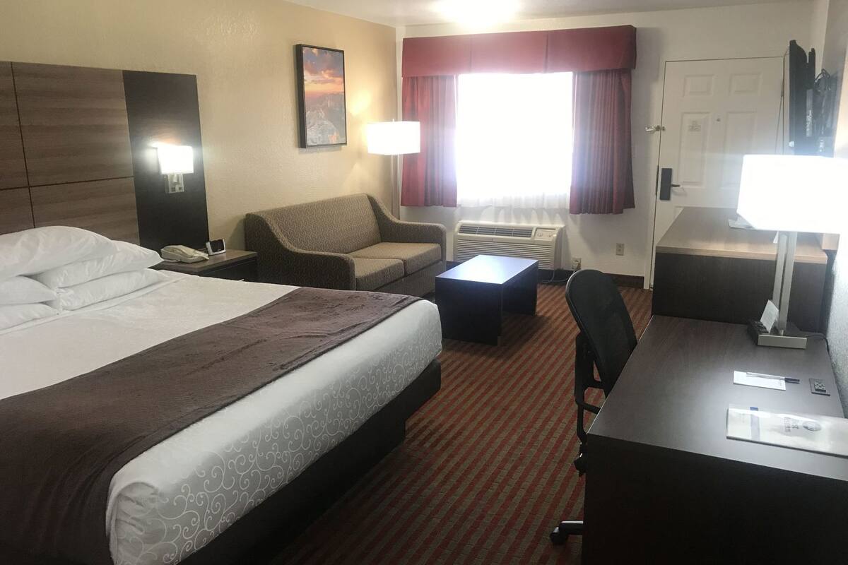 Ein Hotelzimmer mit einem großen Bett, einer Couch, einem Schreibtisch und einem Sessel. Es gibt ein Fenster mit Vorhängen, einen Wand-Fernseher und einen Bilderrahmen.