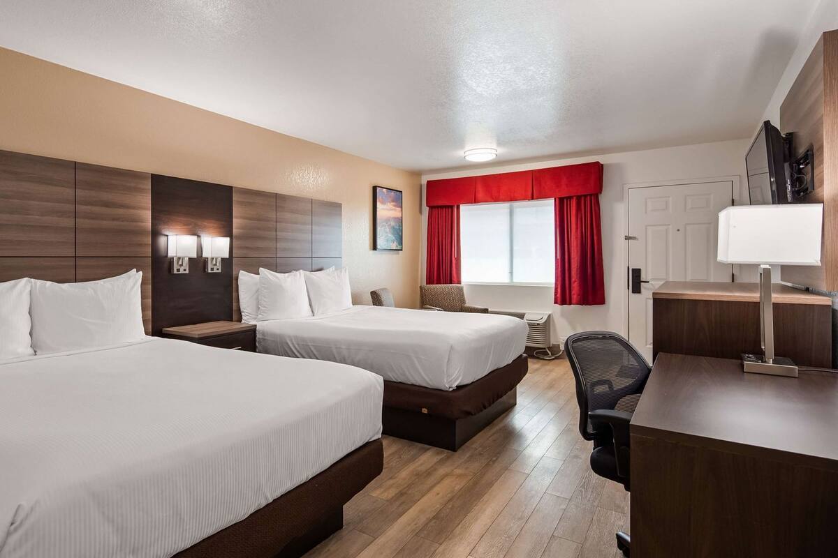 Ein Hotelzimmer mit zwei Betten, einem Schreibtisch, einem Stuhl, einem Fenster mit roten Vorhängen und einem am Wand befestigten Fernseher.