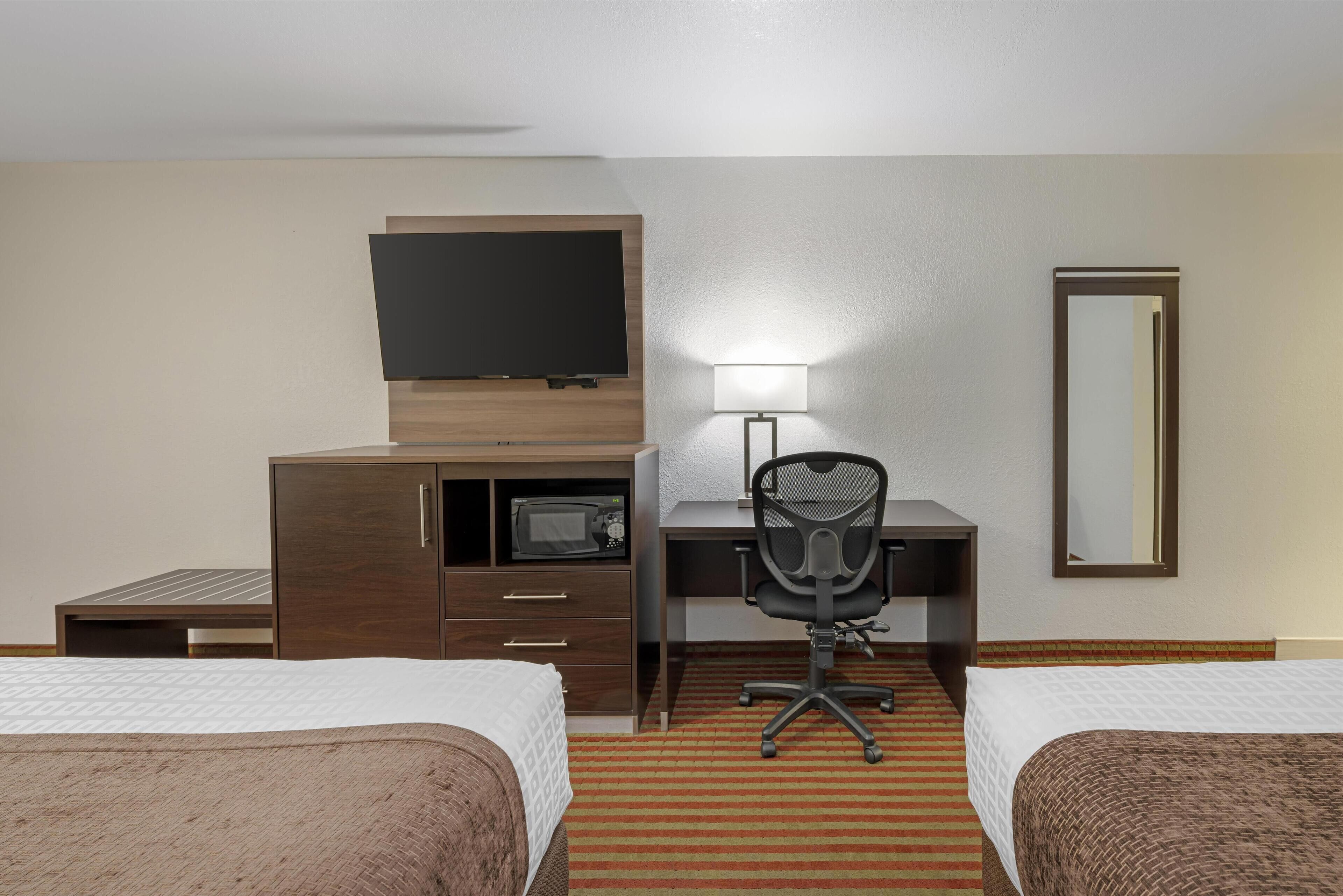 A hotel room with two beds, a desk with a chair, a flat-screen TV, a microwave, a lamp, and a mirror.