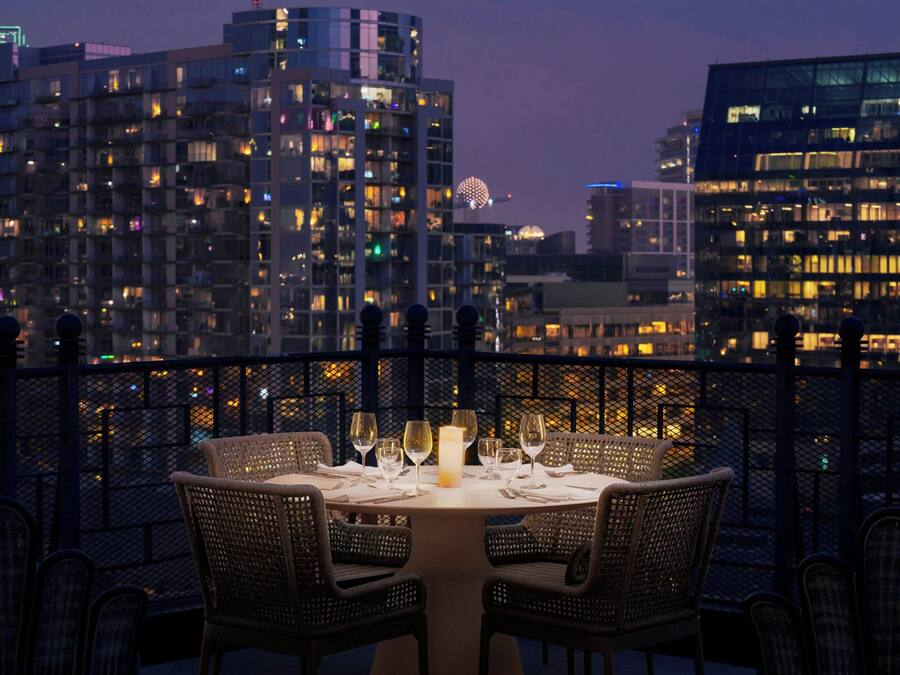 Un comedor con una mesa dispuesta para dos personas, rodeada de sillas de mimbre, con vista a un paisaje urbano al atardecer.