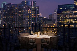 A dining area with a table set for two, surrounded by wicker chairs, overlooking a cityscape at dusk.