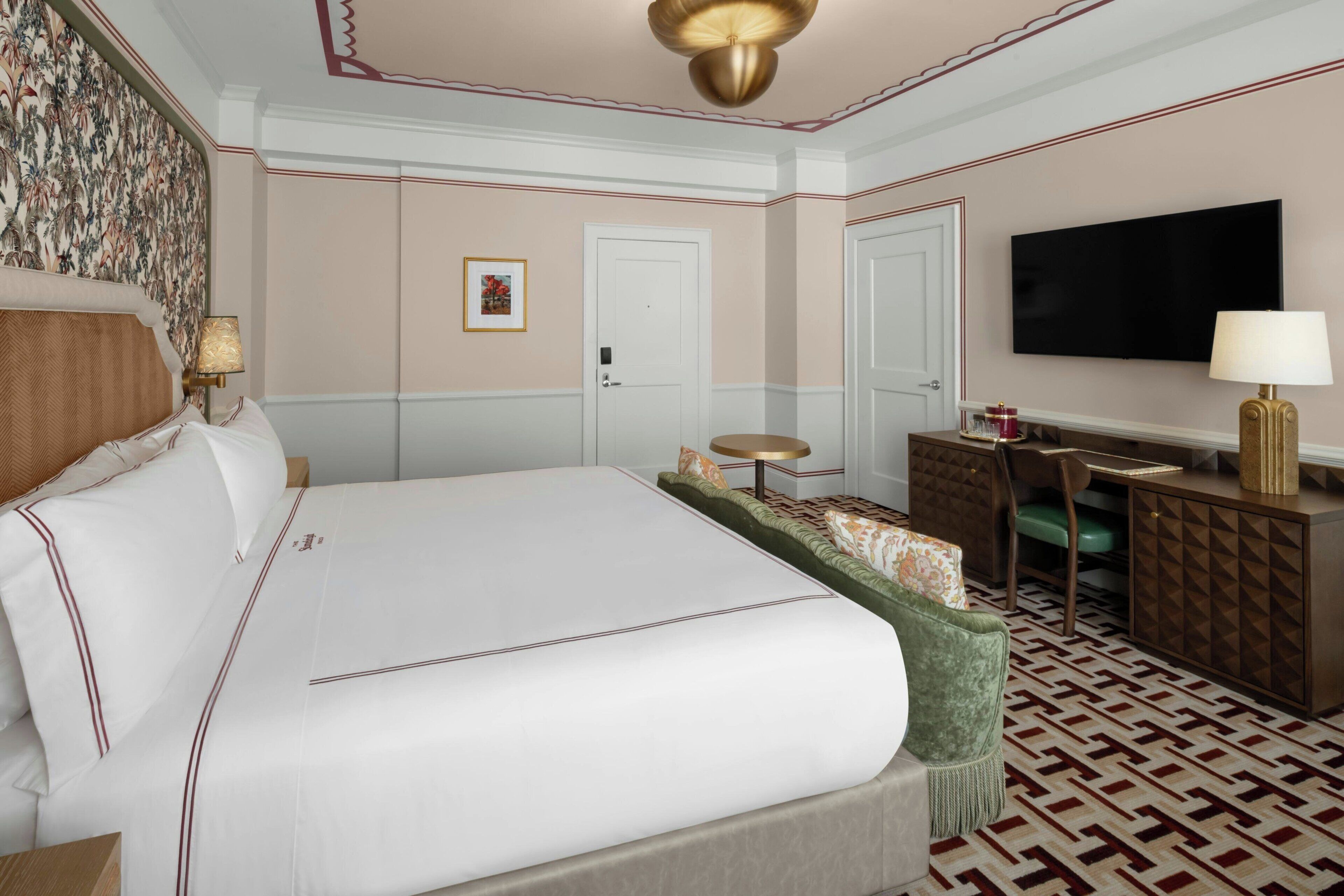 A hotel room with a large bed, a desk, a television, and a patterned carpet.