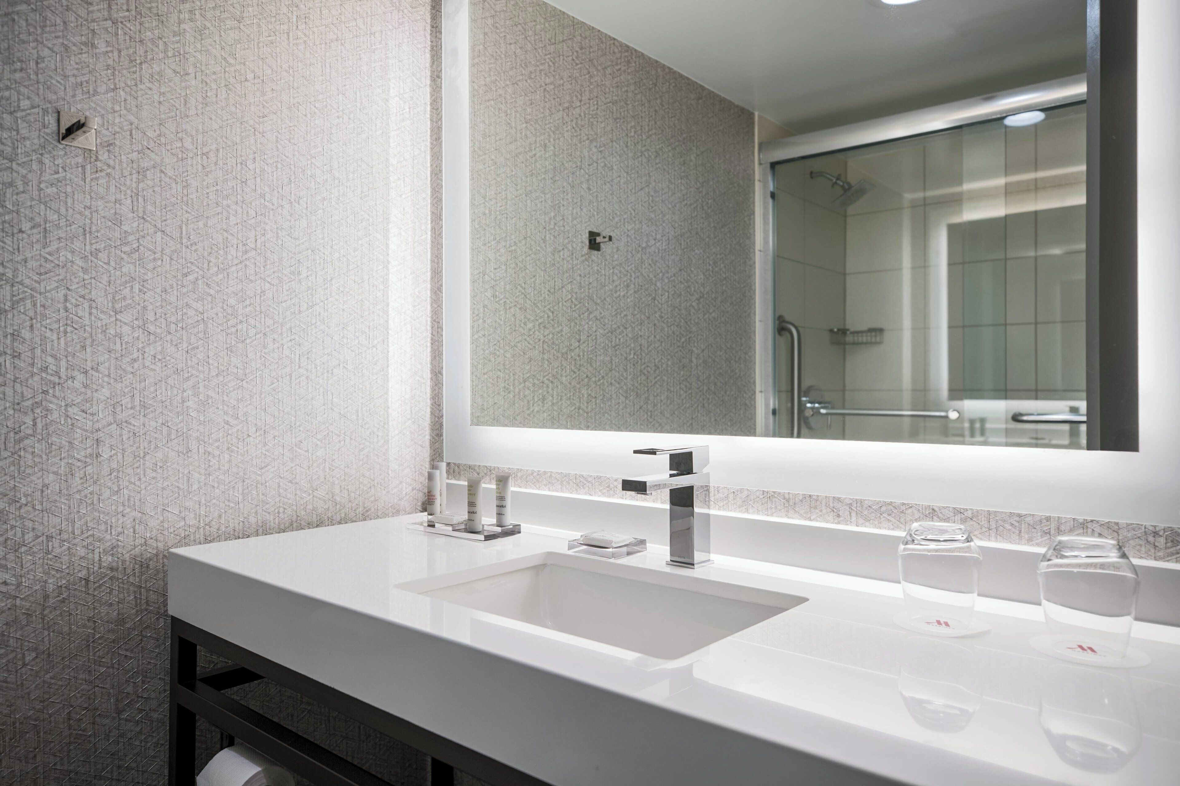 A modern bathroom with a large mirror, two sinks, and a shower visible in the reflection.