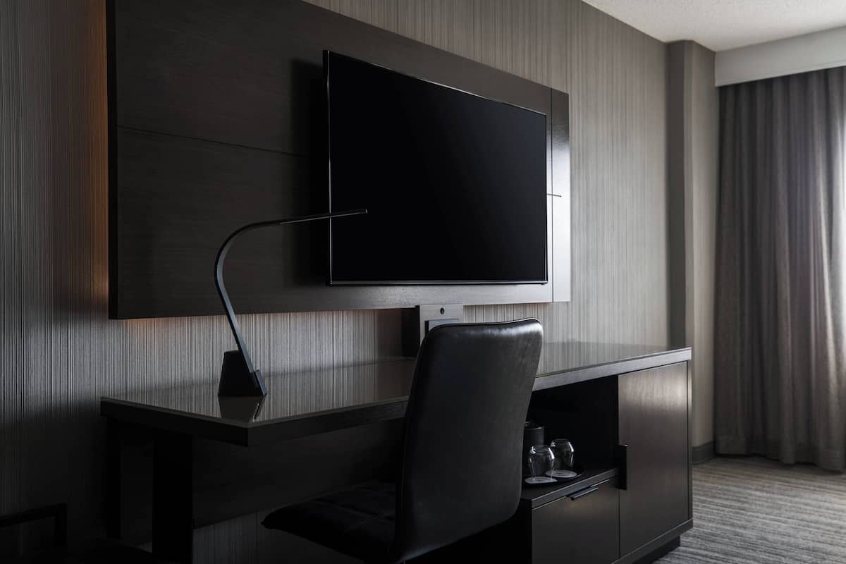 Une chambre d’hôtel moderne dotée d’un téléviseur à écran plat fixé sur un mur en bois foncé, d’un bureau noir élégant avec une chaise, et d’une table à plateau en verre surmontée d’une lampe.