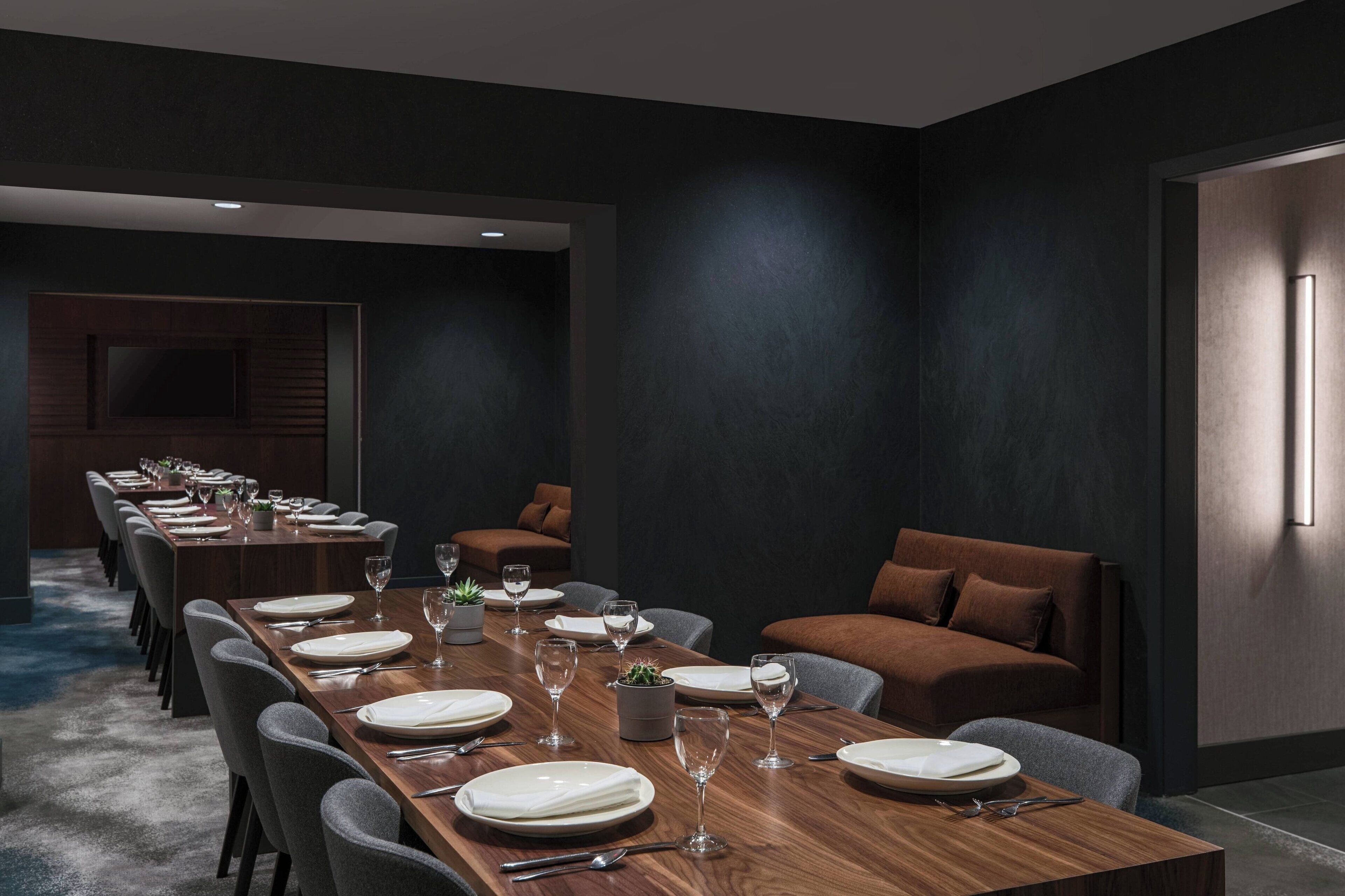A modern dining area with a long wooden table set for a meal, surrounded by grey upholstered chairs.