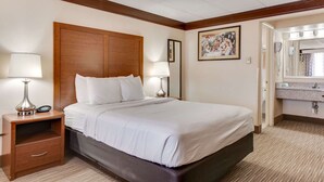 A hotel room with a large bed, two bedside tables with lamps, a wooden headboard, a framed picture on the wall, and a bathroom with a sink and mirror. - Best Western Movie Manor (Monte Vista)