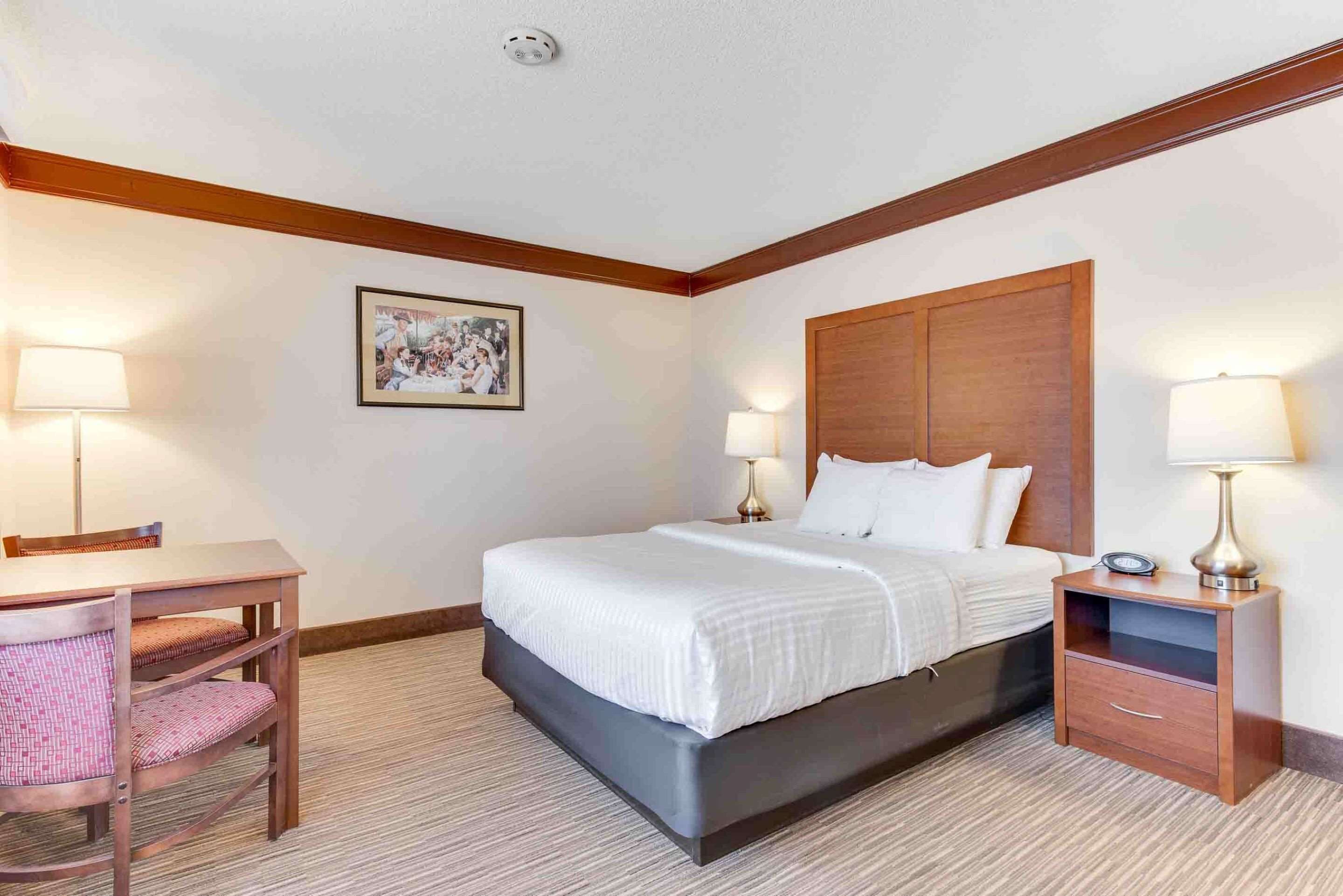 A hotel room with a bed, bedside table, lamp, chair, and a framed picture on the wall.