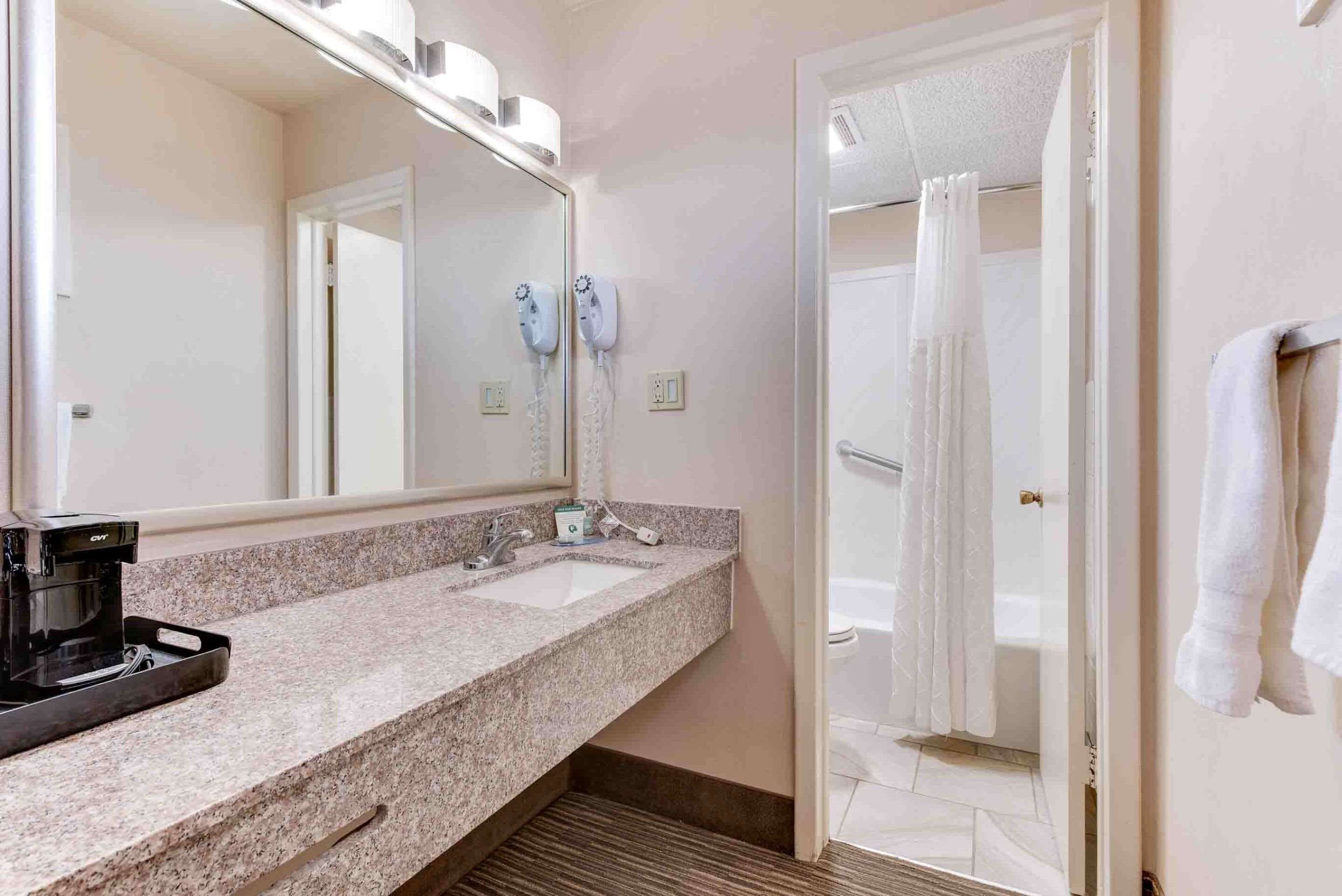 Une salle de bain d’hôtel avec un comptoir en granit, deux lavabos, une machine à café, un grand miroir et une douche avec un rideau.